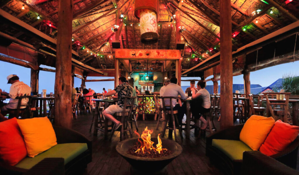 A bar interior at sunset with patrons at tables and a red and green lit roof