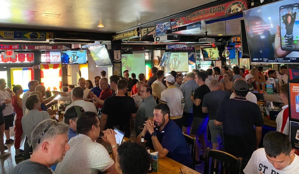 A crowded pub with fans sitting and standing watching soccer on TVs