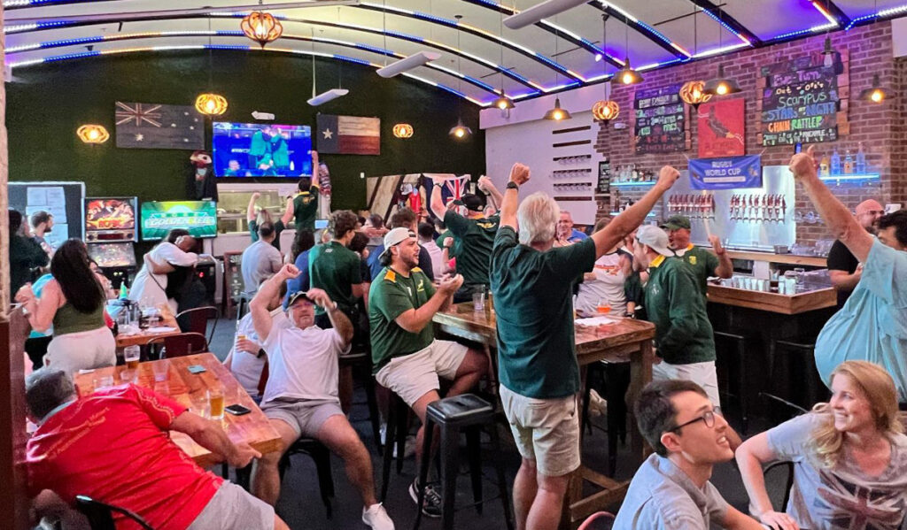 A crowd of people in green shirts celebrate in a brewery taproom