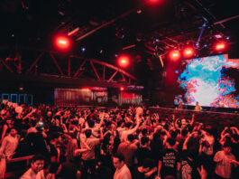 A red-lit nightclub filled with people dancing and a DJ on stage