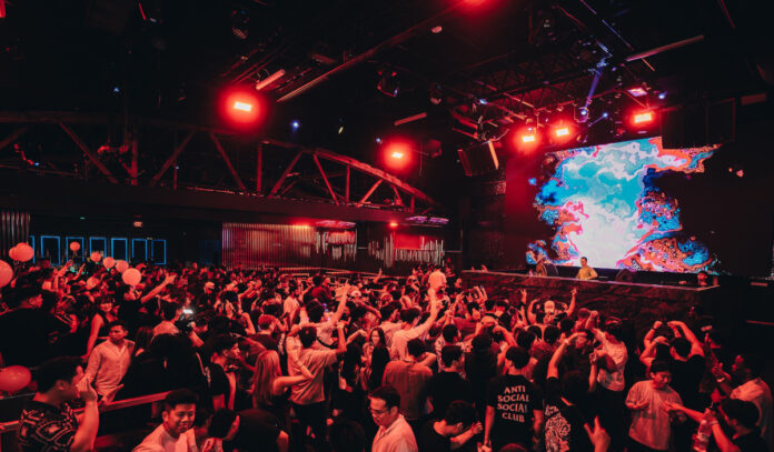 A red-lit nightclub filled with people dancing and a DJ on stage