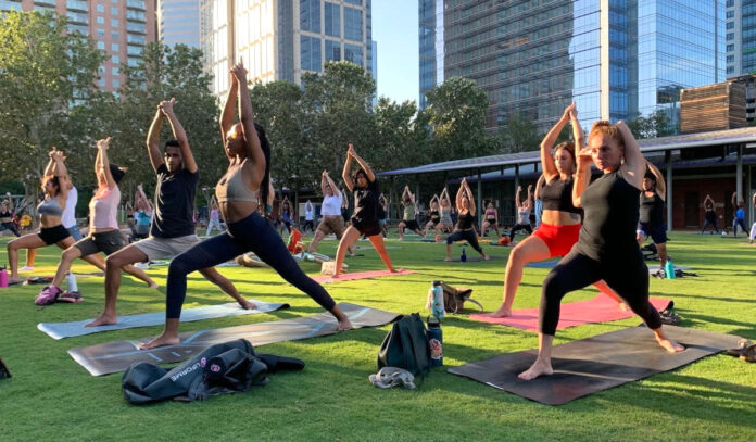 Several people performing a yoga pose in unison on grass near skyscrapers