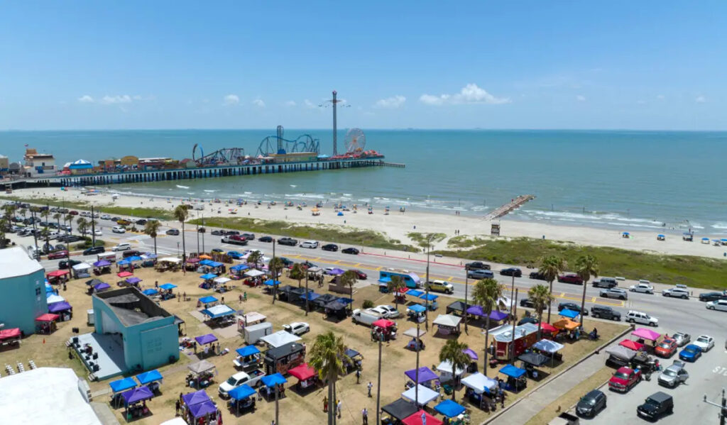 An aerial of a park festival with market stalls next to a beach and a pier in the background