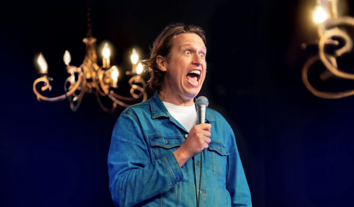 Comedian Pete Holmes performing with a microphone and wide eyes and open mouth