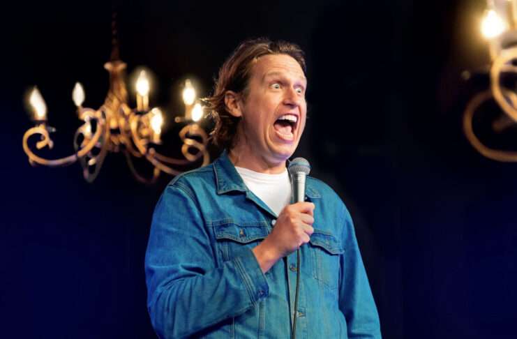 Comedian Pete Holmes performing with a microphone and wide eyes and open mouth