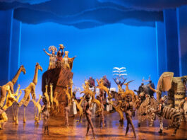 The company of "The Lion King" in animal suits lift their arms to a lion held above them