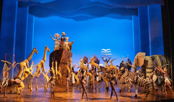 The company of "The Lion King" in animal suits lift their arms to a lion held above them