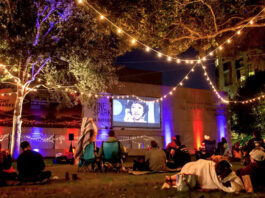 An outdoor movie screening on the side of a building as people watch from blankets and chairs under strings of lights