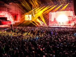 A concert crowd and performers on a stage with yellow beams of light emitting from the stage