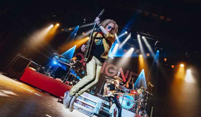 A guitarist jumps in midair while a band plays behind him