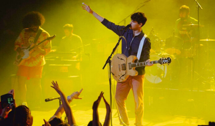 The lead singer of Vampire Weekend stands on a yellow stage with an arm outstretched while performing