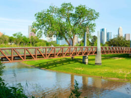 Two people ride bicycles on a bridge that crosses a bayou with a downtown skyline in the background