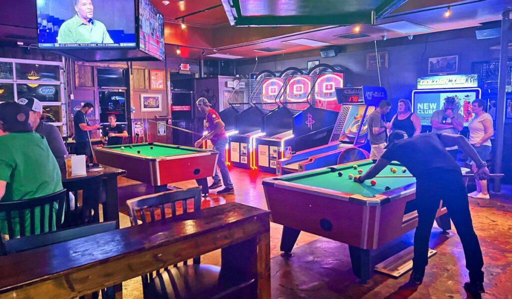 A barroom interior with people playing pool next to other arcade games