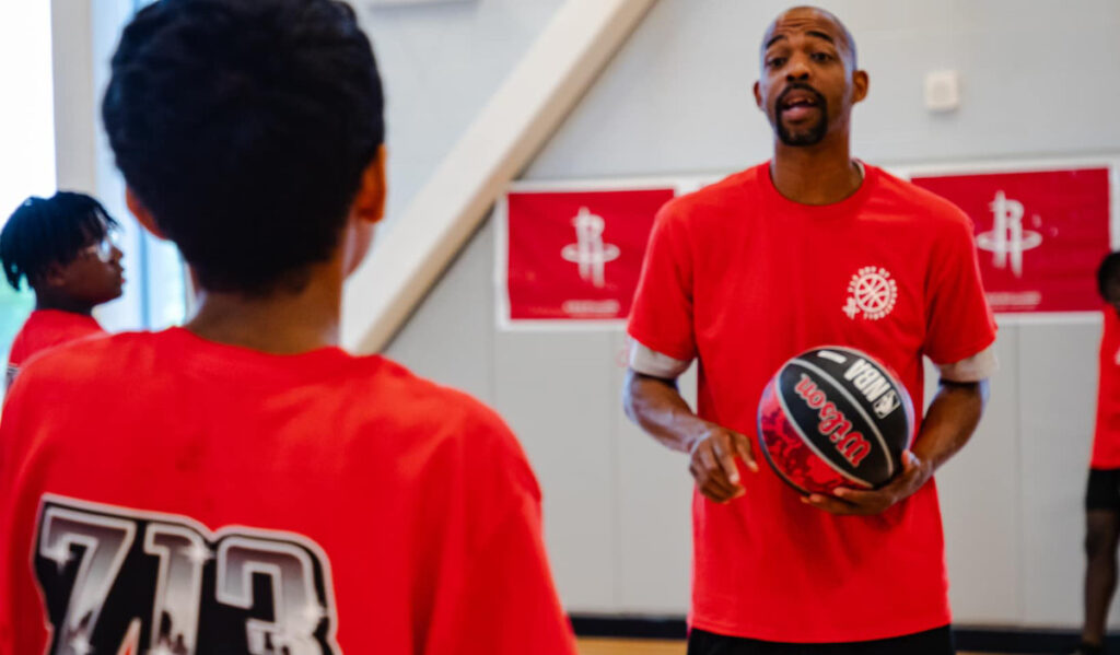 Rafer Alston holding a basketball talking to a child that is facing him, wearing a shirt with "713" on it