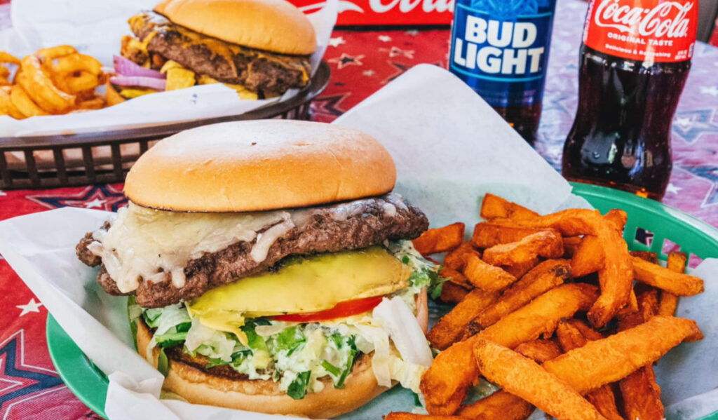 A burger with several toppings and sweet potato fries next to a beer and a coke