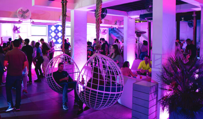 A bar interior lit in purple with two people sitting in suspended swings
