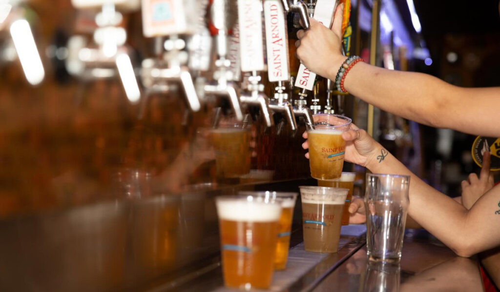 Hands pouring glasses of Saint Arnold beer from a wall of taps