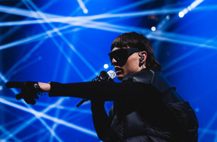 Peso Pluma singing on stage and pointing with a blue laser backdrop