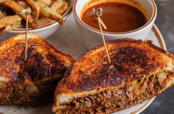 A grilled sandwich cut in halves on a plate with fries and a sauce dip